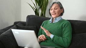 Positive senior mature woman sitting on the couch, laughing and looking at the laptop screen, talking via video call and reacting to a funny joke from her friends, looking on the webcam