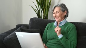 A middle-aged gray-haired lady wearing a green sweater jumper is looking and waving at the laptop screen, grandmother senior lady is talking online, video chatting concept