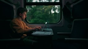 Handsome young man rides in the evening in a train compartment at a table with a laptop and takes photos from the window on a smartphone camera.Guy uses Internet on gadgets while traveling by train.