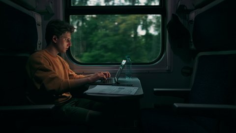 Handsome young man rides in the evening in a train compartment at a table with a laptop and takes photos from the window on a smartphone camera.Guy uses Internet on gadgets while traveling by train.