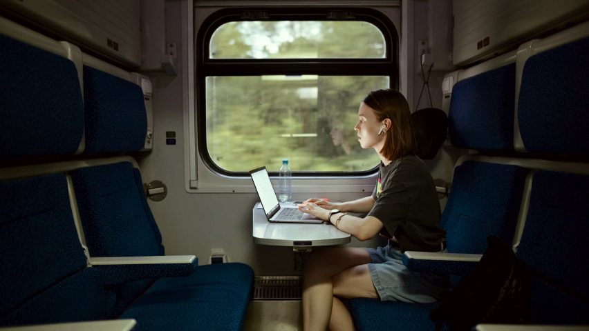 Beautiful young woman in casual clothes working on a laptop at a table in a train and communicates on wireless headphones. Female freelancer working on laptop while traveling by train. Royalty-Free Stock Footage #1067154754