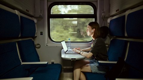 Beautiful young woman in casual clothes working on a laptop at a table in a train and communicates on wireless headphones. Female freelancer working on laptop while traveling by train.
