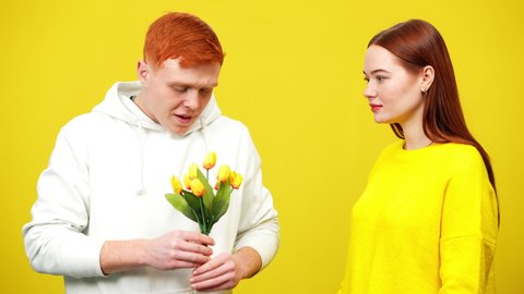 Shy redhead man giving bouquet of yellow tulips to smiling surprised beautiful woman with dental braces. Portrait of timid millennial Caucasian couple at yellow background. Romance and love concept.