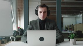 Smiling businessman in headphones using laptop computer for video conference while sitting at office. Handsome man talking on video call. Webcam company team meeting concept.