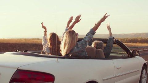 Group of friends ride in cabriolet car waving their hands from side to side, enjoying life, rear view. Friends in cabriolet car have fun wanderlust