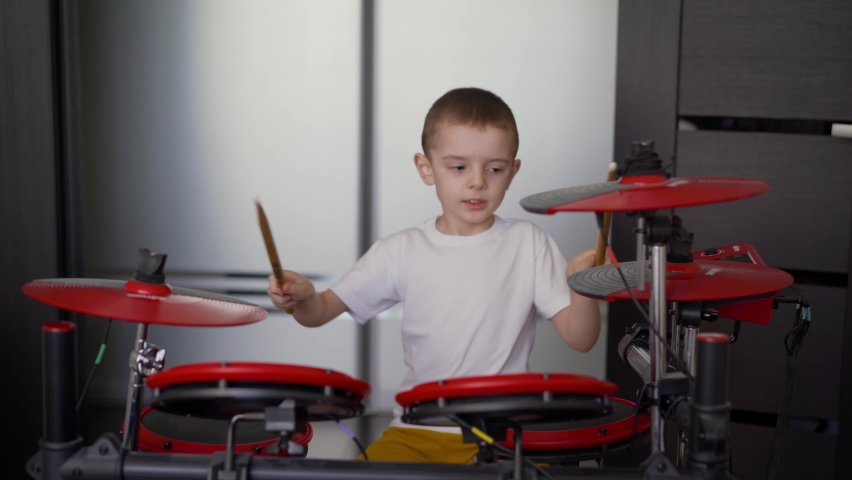 A boy plays electronic drums at home. A little boy enjoys playing the electronic, red, drum at home. The boy at home on self-isolation exercises drums.