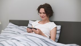 Happy young woman wears white t-shirt lying in bed rest relax spend time in bedroom home in own room hold in hands use mobile cell phone watching tv show film social network video. Real estate concept