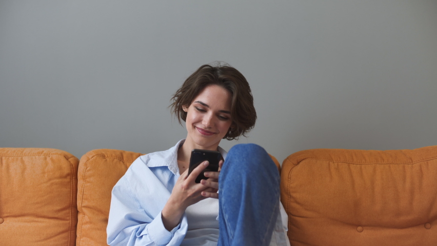 Smiling young woman in shirt sit on sofa couch rest relax spend time in living room lounge home in own house hold using mobile cell phone chatting serfing browsing typing sms dream Real estate concept Royalty-Free Stock Footage #1067175037