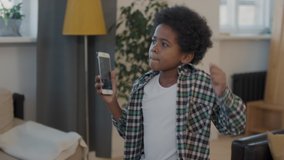 Waist-up shot of cute and stylish african boy dancing to music in living room with smartphone in hands filming funny videos