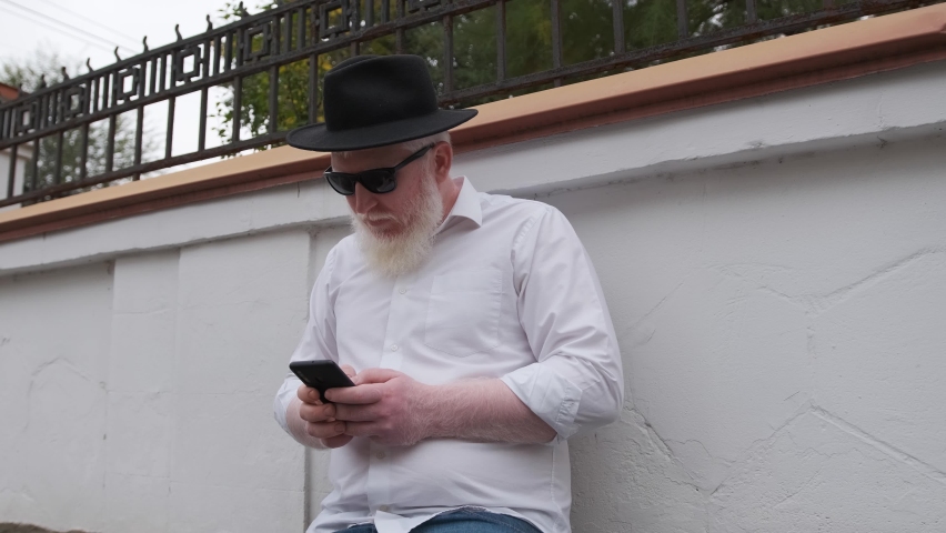 Old man with albinism browsing on the internet while sitting next to his house, man wearing a black hat and sun glasses to protect himself.