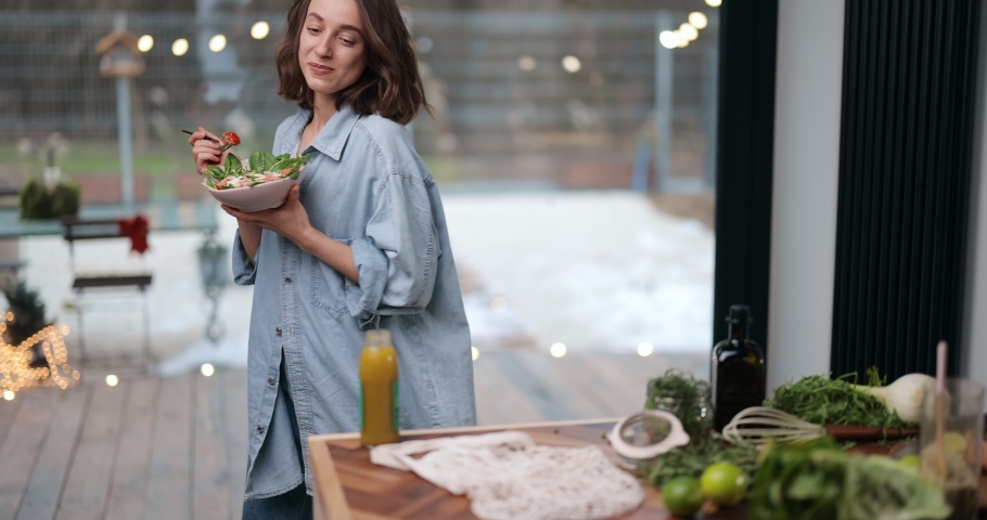 Beautiful young woman eating healthy salad at cozy living room near the window at home, dancing and feeling easy. Healthy veggie food and wellness concept. Royalty-Free Stock Footage #1067178580