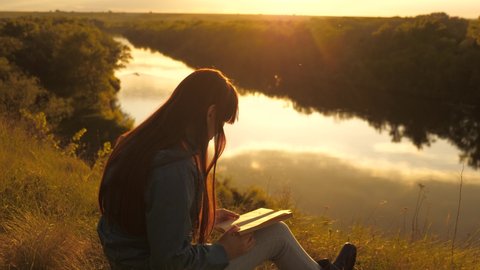 Girl traveler reads a book at a halt. Young woman student with textbook outdoors. A young girl sitting on a mountain on a green lawn by the river preparing for an exam in the park.
