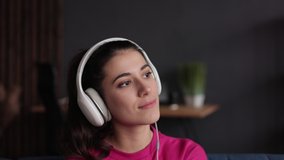 Close up portrait female listening to music in modern headphones. Pretty young woman dancing and listening music with white headphones. Beautiful girl enjoying music on couch at home.