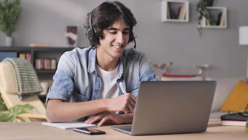 smiling young man in headset working on a project in video chat conference using laptop at home,male student at his desk talking at the computer with a virtual coach teacher,taking notes Royalty-Free Stock Footage #1067185996