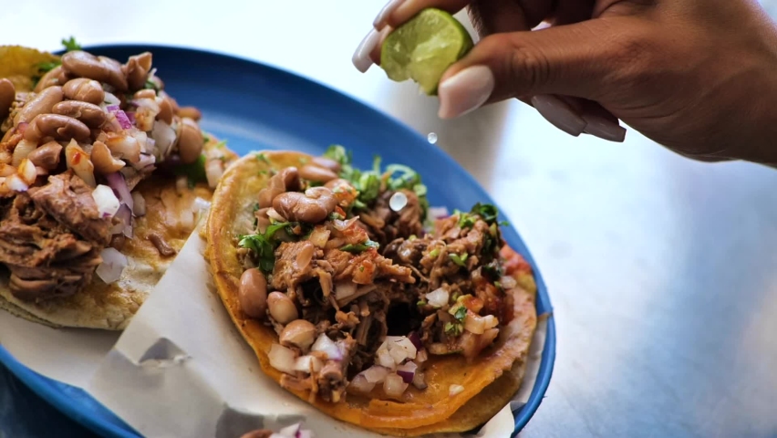Lime juice is dripped onto a serving of two delicious Mexican tacos, complete with beef, beans and cilantro. Close up in slow motion. Royalty-Free Stock Footage #1067190652