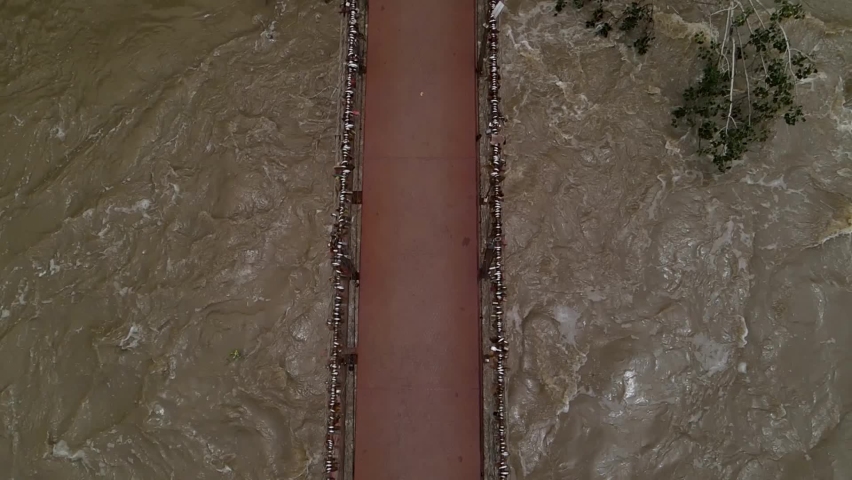 Elderly man and woman standing on a suspension bridge, looking at a strong flowing river below the bridge. Drone footage in slow motion, top down to reveal bridge, river, and people. 1080p 30 fps.