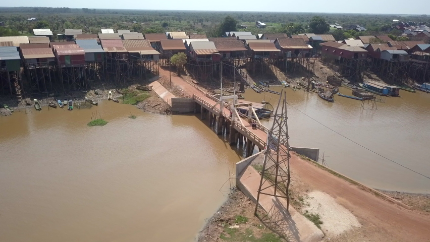 Drone view of a long tail fishing boat passing under a small bridge. Floating village along the banks of river as backdrop. 	
