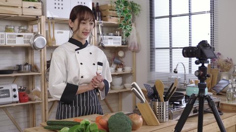 beautiful asian culinary specialist vlogging at home. introducing ingredients with gestures before cooking a meal. holding cucumber and onion in front of digital camera.