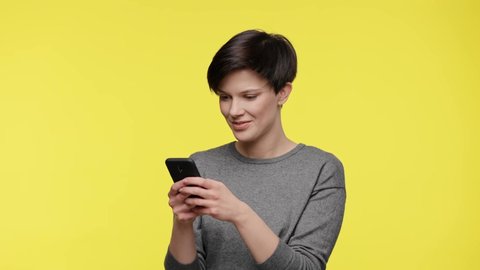 Color of the year 2021. Ultimate Gray and illuminating. Surprised young beautiful woman in gray sweatshirt using smartphone, get shocking and unbelievable news, isolated on yellow background