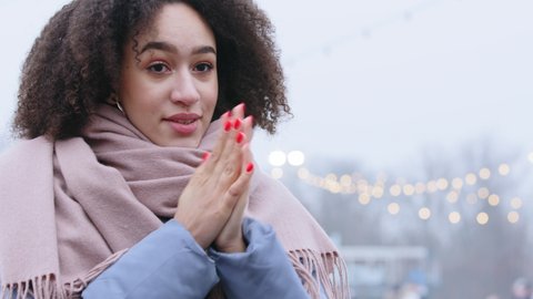 Portrait of afro american woman with curly hair wears warm clothes jacket and scarf stands alone in cold weather in city warms her hands rubs palms blowing hot air on fingers with perfect manicure