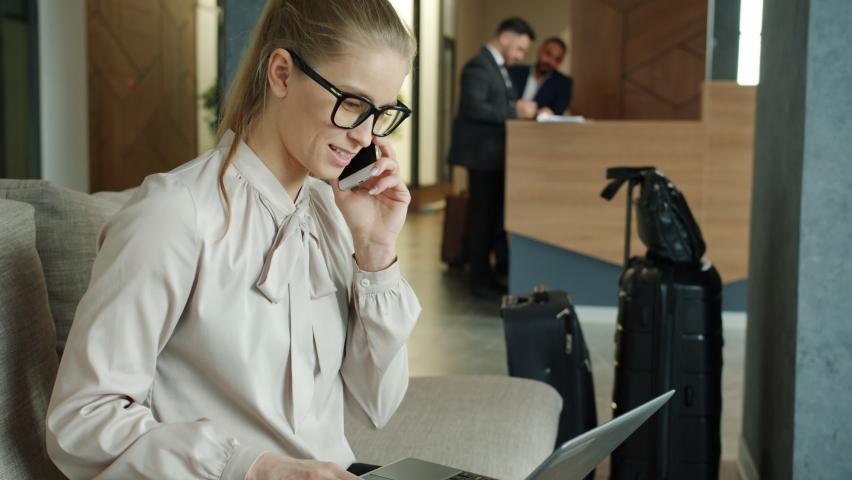 Cheerful businesswoman talking on mobile phone and working with laptop in hotel hall sitting on coach concentrated on business communication smiling | Shutterstock HD Video #1067213302