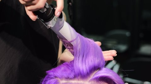 A woman sits in a beauty salon with pink-purple hair, the master makes her large curls with a curling iron  