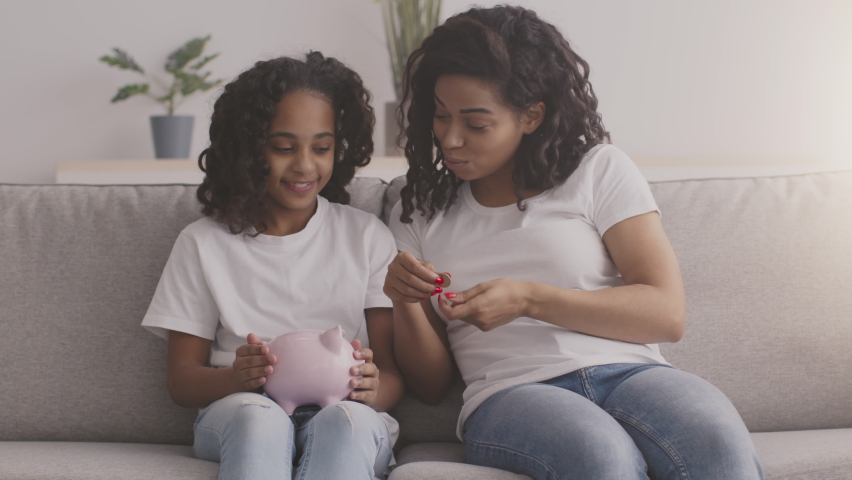 Financial education. Young black mother putting money into daughters piggybank and talking about financial literacy at home, slow motion | Shutterstock HD Video #1067214340