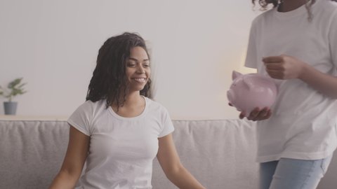 Financial responsibility. Cute black girl sitting on mothers knees and putting coin into piggy bank, slow motion