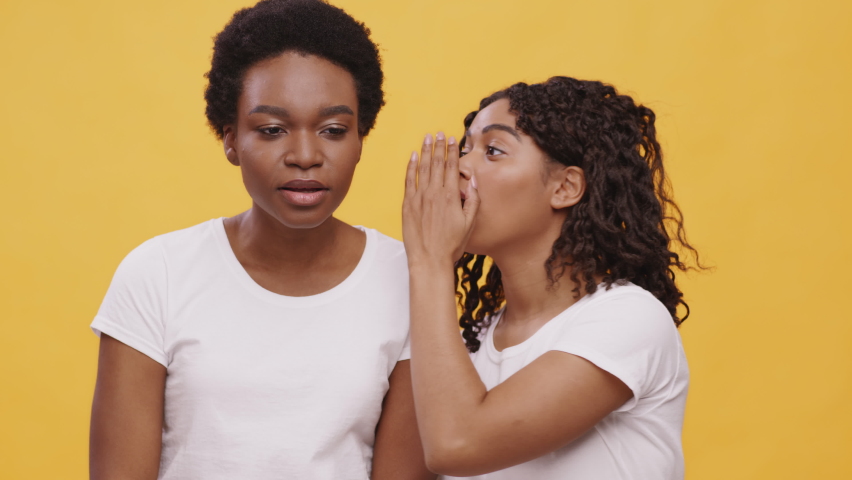 Female gossips. Young african american woman whispering gossip or telling confident information to her shocked black friend, orange studio background. Unexpected or unpleasant news | Shutterstock HD Video #1067214517