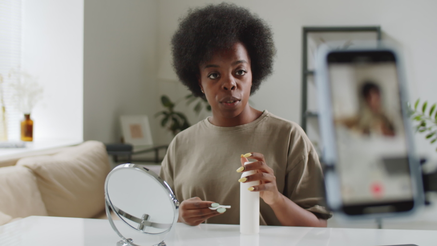 Young beautiful Afro-American woman sitting at table in living room, looking at smartphone camera and showing how to apply lotion to facial skin with cotton pad while recording video for beauty vlog | Shutterstock HD Video #1067226175