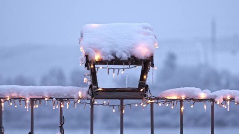 Bird feeder covered by snow with christmas lights during snow fall. Small wooden house for hungry birds. Winter and cold weather.