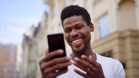 Closeup surprised man talking on video from smartphone in city. African american man making hand gesture outdoors. Portrait of excited afro guy having video call on street.