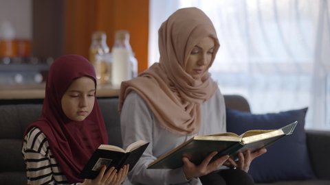 Muslim mother and daughter with a headscarf read the Quran together. Muslim mother and daughter sit next to each other at home and read the Quran. The Quran reading concept.