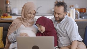 The Muslim woman wearing a headscarf, her daughter and her husband are happy while video chatting on the laptop and showing her belly for the baby to be born. New sibling concept. Close up.