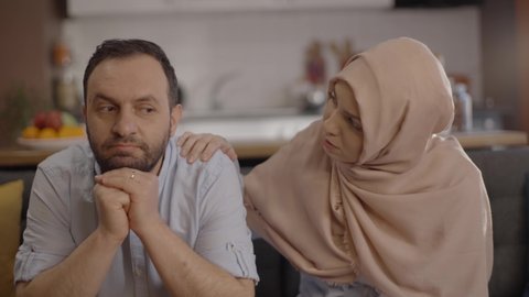  The Muslim woman wearing a headscarf trying to cheer her up by touching her sad husband's shoulder, speaks to her husband, hugs him, comfort him.