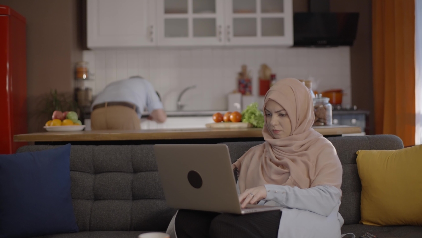 A young woman in hijab, working from home, describes what she should do to her husband, who prepares a meal in the kitchen. Remote working concept. Royalty-Free Stock Footage #1067247295