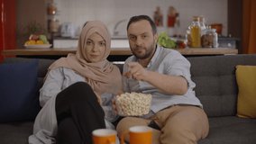 Muslim husband and wife watching a movie while eating popcorn, get excited, pull the popcorn between them and laugh.