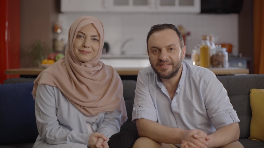 The bearded man and his wife in hijab, making a video call in their house, greet their loved ones and speak. Image through the eyes of the computer camera. 
