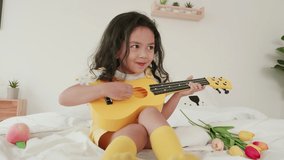 Kid girl in a yellow blouse is enjoying a joyful playing guitar on the bed