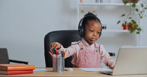 Concentrated afro american little girl child sitting at home at table draws picture chooses pencil wears headphones listens to Internet teacher watching video lesson on laptop online conference chat