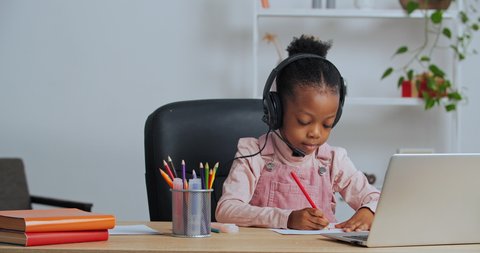Concentrated afro american little girl child sitting at home at table draws picture chooses pencil wears headphones listens to Internet teacher watching video lesson on laptop online conference chat