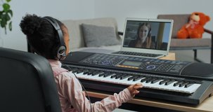 View from behind small schoolgirl african american child in headphones looks into laptop speaks online chat learn to remotely play piano synthesizer keyboard instrument with help of internet teacher