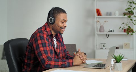 Afro american student guy in casual shirt in cage wears headphones speaks into laptop webcam with teacher answers exam, freelance business man conducts interview negotiation online video conference