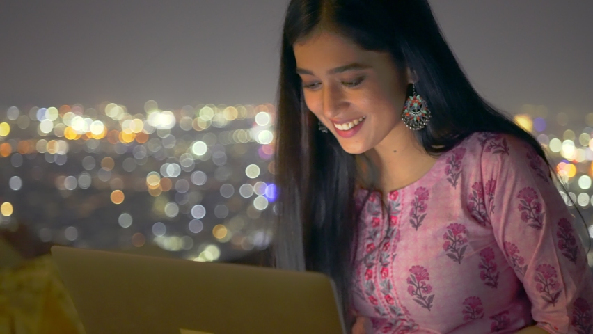 Shot of a cheerful and attractive young Indian female sitting on a terrace of a building is drafting an email or doing office work using a laptop computer against the illuminated cityscape at night | Shutterstock HD Video #1067269267