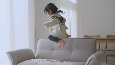 Active Asian toddler girl jumping on the couch in the living room at home. Preschool child holding a stuffed dog in her arm, bouncing on the sofa, playing alone at home. 2 to 3 years old