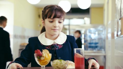 A girl in a beautiful school uniform carries a tray with a delicious and healthy lunch. Other children choose their meals at the cash register in the school cafeteria