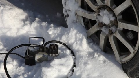 Man hand inflates with tire inflator hose portable electric air compressor the car wheel on white snow background at Sunny winter day, vehicle tyres pressure control, tyres self-repair