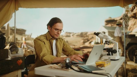 Archaeological Digging Site: Famous Male Archaeologist Doing Research, Using Laptop, Analysing Fossil Remains, Ancient Civilization Culture Artifacts. Team of Historians work on Excavation Site