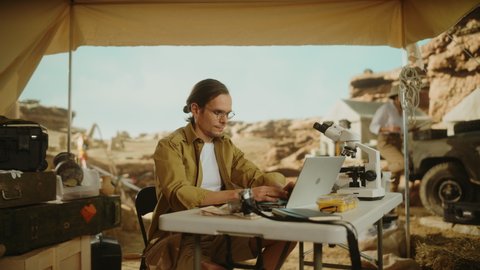 Archeological Digging Site: Portrait of Great Male Archaeologist Doing Research, Using Laptop, Looks at the Camera Smilingly while Holding Fossil Remains or Ancient Civilization Culture Artifacts