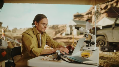Archaeological Digging Site: Smart Male Archaeologist Doing Research, Using Laptop, Analysing Fossil Remains, Ancient Civilization Culture Artifacts. Team of Historians work on Excavation Site